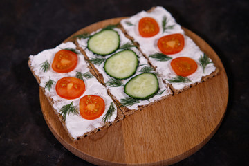 Crispbread with curd cheese and vegetables on a wooden board, on a dark background. Space for text. Flat lay, top view. Diet snack.