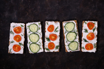 A set of crispbread with curd cheese and fresh vegetables, cherry tomato and cucumber, on a dark background. Space for text. Flat lay, top view. Diet snack.