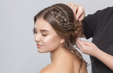 Naklejka premium Portrait of a beautiful sensual light brown haired woman with a wedding hairstyle in a beauty salon. The hairdresser does the hairstyle. Wedding hairstyle.