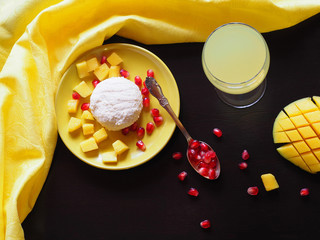 Cottage cheese in a yellow plate, spoon, pomegranate seeds, mango slices, juice in a wine glass and a yellow napkin on a black background, top view.