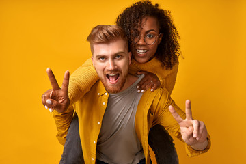 Photo of cheerful cute charming positive nice diverse couple showing you victory sign and giving a piggyback ride to joyful, wearing in monochrome orange, isolated over an orange color background.