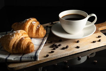 black cofffee in white cup and wheat flour sprinkle croissant on towel on board wood with black background