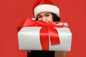Girl in a black dress and Santa hat hiding behind Christmas giftbox of white color with red ribbon.