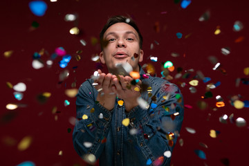 Man in a blue jeans shirt celebrating Christmas and New Year with confetti over red background.