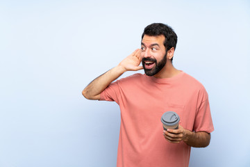 Young man with beard holding a take away coffee over isolated blue background listening to something by putting hand on the ear