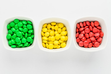 Three squared bowls with small red, yellow and green coated chocolate candies in a squared bowl isolated on white background, top view or flat lay of sweets