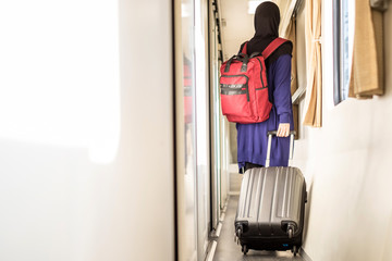 Selective focus of silver suitcase dragged by young muslim traveller wearing hijab and with red backpack in walkway inside train.