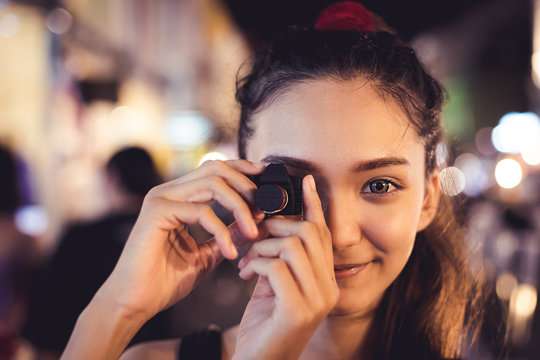 Fototapeta Girl holding small black cemara look pretty to pretend to take a shot, one big eye with smiling face, outdoot at night have some bokeh of light on background
