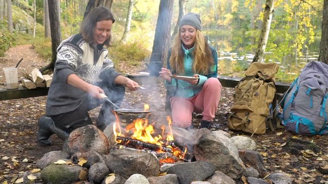 Female friends cooking traditional pancakes over an open fire in the camp outdoor, enjoying the silence and fresh air during a hike in Finland