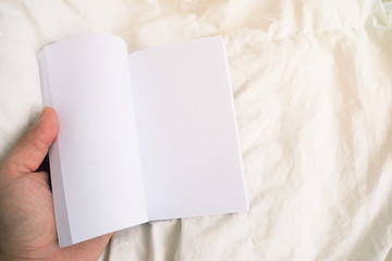 Top view of Hand holding a book with blank white pages with on bed with copy space for add text and notice for presentation and marketing purpose. Media and reading lifestyle concept