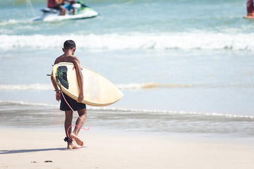 Black skin kid walking with surfboard in a hand to the sea to play surf in sunny day with some wave...