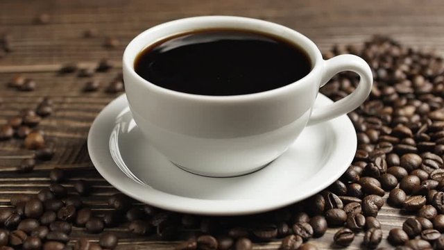 On a wooden table is a white mug of coffee and coffee beans are lying around, close-up drops of coffee falling into a cup.
