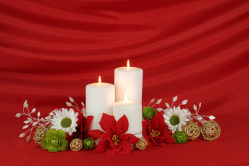 White Christmas candles surrounded by red poinsettia flowers, green roses, white daisies and gold twig balls on a red draped satin background