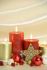 Red and gold burning Christmas candles surrounded by natural potpourri elements and gold stars on a cream satin draped background. Vertical