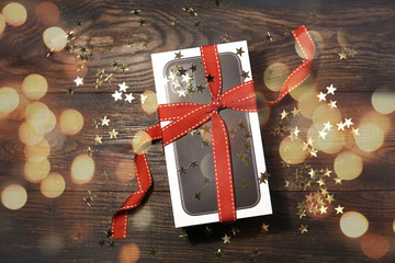 A box of new black Mobile Phone  with red ribbon, lights, golden stars - a gift on wooden table. Top view. Christmas, new year concept    