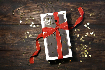 A box of new black Mobile Phone  with red ribbon, lights, golden stars - a gift on wooden table. Top view. Christmas, new year concept    