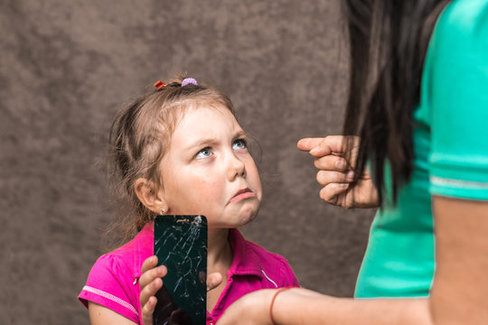 The child accidentally broke the phone and cries. Mom punishes her daughter for a broken phone screen. A little girl holds a cracked phone in her hand and looks at her mother plaintively.