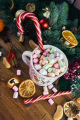 Christmas cup with marshmallows and caramel cane. Wooden background with fir branches, oranges and chocolate chip cookies.