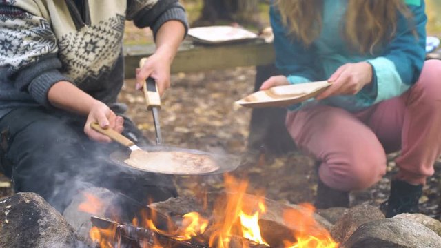 Female friends cooking traditional pancakes over an open fire in the camp outdoor, enjoying the silence and fresh air during a hike in Finland