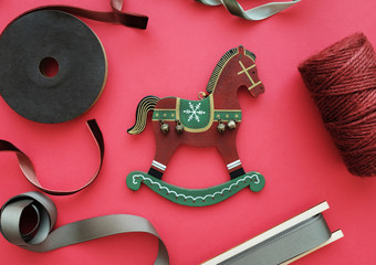 Christmas concept. Top view of little wooden toy horse as a christmas decoration with thread and ribbon bobbins of green, maroon and olive color on the red background. Central composition. Flat lay.