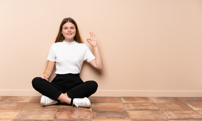 Ukrainian teenager girl sitting on the floor surprised and showing ok sign