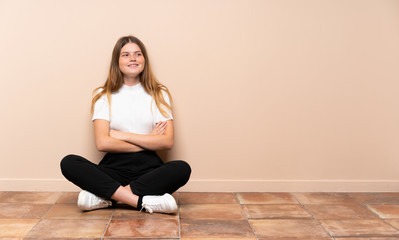Ukrainian teenager girl sitting on the floor with arms crossed and happy