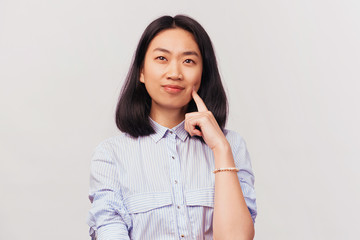 Frowning woman holding forefinger on cheek, looks up. Businesslike beautiful young woman of Asian appearance with black hair dressed in striped office shirt stands isolated white background in Studio.