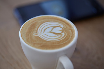 Art in coffee cup. Close up over view of art on coffee toppings . Close up and Selective focus of coffee cup on wood table in front of blur smart phone.