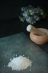 Sea salt is on a black stone table. Nearby is a mortar with a pestle and a bouquet of wildflowers.Still life. A healthy lifestyle. Space for text.