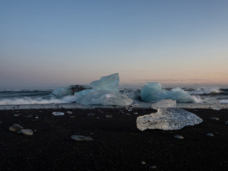 Waves rolling on an iceberg in the mesmerizing Diamond Beach. Some small ice pieces washed up on shore and melt in the sand in the foreground. At the dusk. Breiðamerkursandur, Iceland.