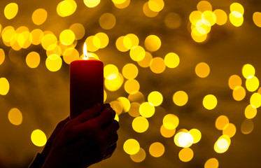 A woman is holding a red candle in her hands. In the background, lights with festive bokeh light...