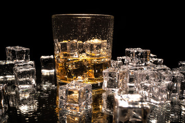 Drinking glass with drink and ice cubes, and more ice cubes on a table, with dark background