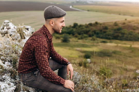 Portrait of pensive man in shirt, cap and suspenders, who sits on the rock top of the hill