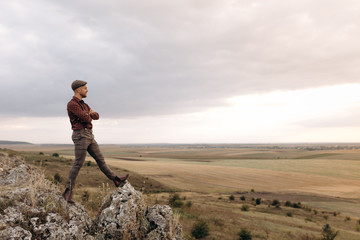 Fototapeta premium Image of a young farmer who stays on the top of the rock hill and looks into the distance to the beautiful landscape of own fields, thinking about future plans. Optimistic mood