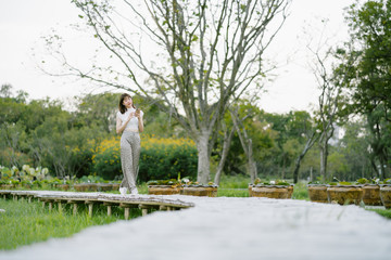 Obraz premium young smiling woman in white clothes with earphones using mobile phone listening to music with her eyes looking at the screen enjoying her moment while strolling on wooden walkway in the park