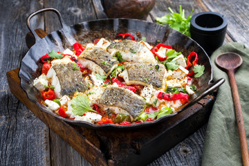 Traditional fried Greek cod fish filet casserole with feta cheese and vegetable closeup view in a rustic wrought iron pan on a wooden board