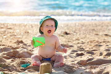 Happy baby in a cap sits on the beach and holds in his hand a green bucket at sunset