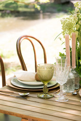 Festive table decor in openair next to the pond. Green color with wooden cutlery. Different natural white flowers. Wooden table and chairs. Wine glasses. Luxury wedding, party, birthday.