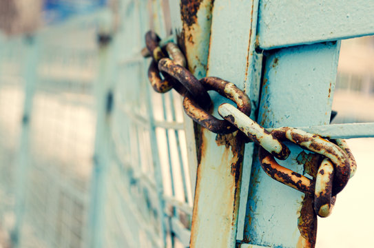 Old rickety iron gates of blue color closed on an old rusty chain. Blurred background, bokeh, grunge tinted. The concept of outdated protection, weak security.