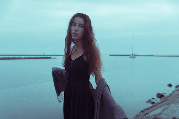 A beautiful and sad girl stands on a stone pier with a landscape on the sea. it rains