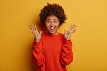 Half length shot of optimistic curly haired woman keeps palms raised, dressed in red jumper, feels...
