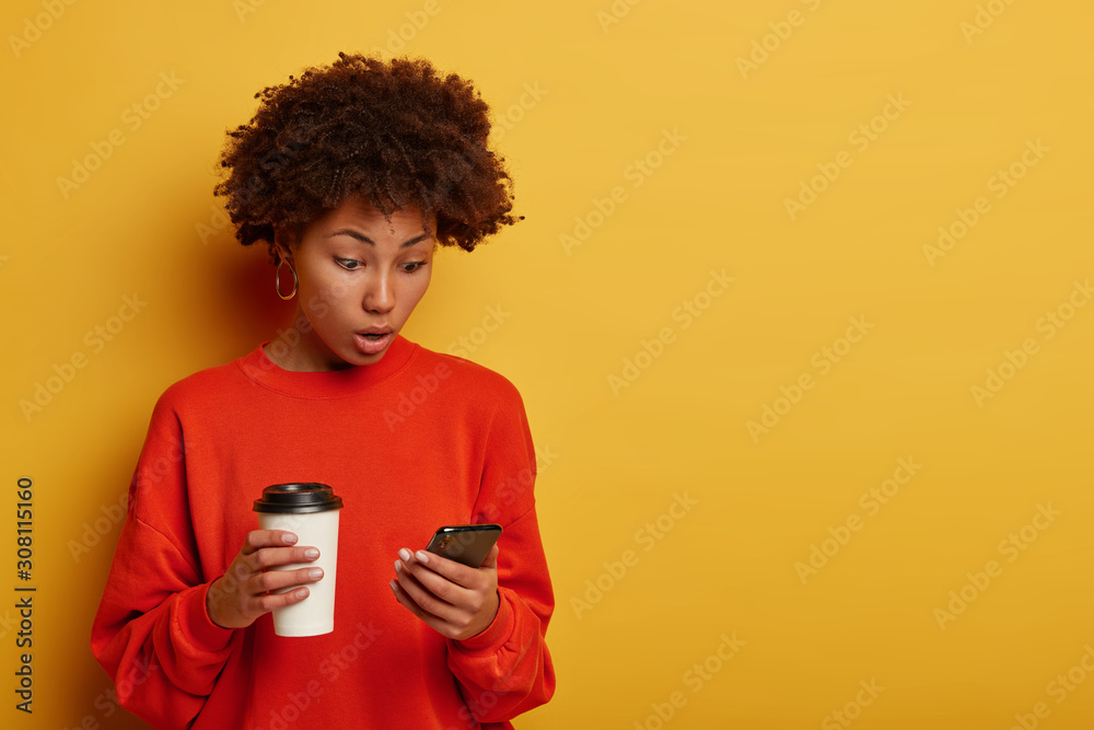 Wall mural Photo of surprised dark skinned woman gasps from surprisement, shocked to see new post, connected to wireless internet, reads astonishing news in internet, drinks coffee to go, watches video