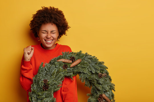 Yeah, holiday is coming. Cheerful curly woman clenches fist, anticipates miracle happen, holds beautiful Christmas wreath, dressed in bright jumper, isolated on yellow wall, free space aside