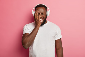 Joyous dark skinned plump man laughs out from positive emotions, covers face with hand, relaxes with music, wears modern stereo headphones, has fun, isolated on pink wall, stands with eyes closed