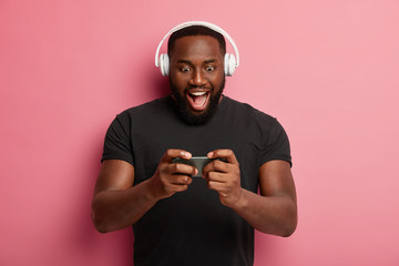 Excited bearded man holds modern smartphone horizontally, plays online game, uses heeadset, stuck at gadget screen, dressed in black t shirt, has fun alone, isolated on pink wall, has free time