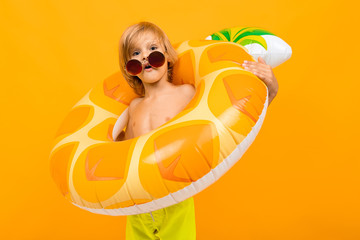 portrait of a fun european blond boy in yellow swimming trunks and sunglasses with a swimming circle pineapple on an orange wall