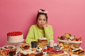 Upset gloomy woman wants to eat sweets and confectionery, poses at table served with many desserts,...