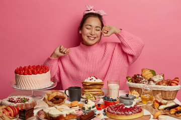 Happy Asian birthday girl comes on tea party, eats sweet delicious cakes, streches hands with enjoyment, surrounded by many desserts, poses against pink background. Sweet tooth, calories concept