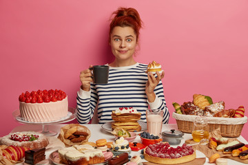 Ginger girl sits at festive table overloaded with many sweet desserts, eats yummy freshed baked...
