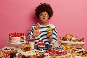 Serious displeased woman with Afro hairstyle shows refusal gesture, holds croissant, denies eating...